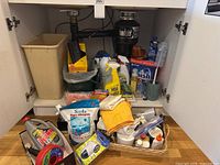 Under-sink cabinet showing garbage bin, compost pail, multiple cleaning bottles, sponges, cloths and basket
