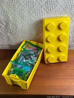 Closed yellow Lego-style storage bin beside open base filled with bagged bricks