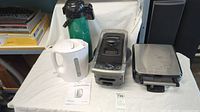 Four small kitchen appliances: green airpot, white kettle, black/silver deep fryer, stainless waffle maker