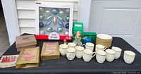 Overview of all items on table including Noritake cups, saucers, several boxed ornament sets, individual ornaments