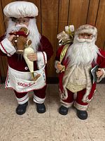 Both Santa figures standing side by side against wood panel wall