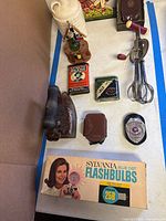 Group view showing flashbulb box, Weston case, cast iron iron, tins, badge, whisk, ceramic bottle and figurine