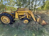 Full left side view of yellow tractor with bucket, showing rust, vegetation and overall condition