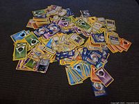 Pile of assorted Pokémon cards spread on dark surface, showing backs and mix of energy and trainer fronts