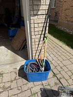 Blue tote beside garage containing assorted garden tools and spade leaning on wall