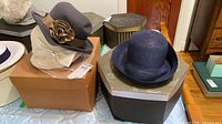 Two hats on boxes: dark gray felt hat with ribbon rosette on Steacy's box, navy woven straw hat on Steacy's box