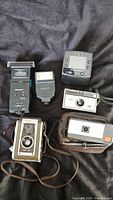Group shot of six items: Duaflex II camera, two flash units, compact 35 mm camera, grey point-and-shoot camera in case, square digital camera body
