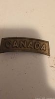 Front of brass CANADA shoulder title pin showing raised lettering