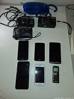 Group shot of all items: five smartphones, three film cameras, blue landline phone, Olympus voice recorder