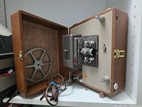 Projector in open case showing reel arm, lens, power cord