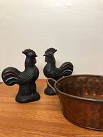 Copper bowl and two wrought iron rooster figurines on wood surface