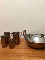 Four copper measuring cups and two nesting copper/stainless bowls on wood surface