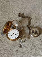 Gold-tone pocket watch open to show dial and case interior beside silver-tone watch and chain