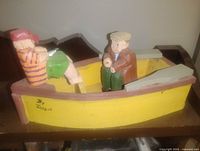 Starboard view of yellow wooden boat with two carved fishermen, signature visible