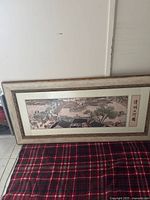 Entire framed embroidery shown leaning against wall on plaid cloth