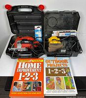 Open cases showing jigsaw with cord, sealed blades, sander, sanding sheets, two DIY books