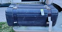 Overall view of black plastic storage trunk showing size, surface wear, latches, missing straps