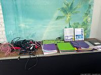 Cables piled left, two keyboards, stacks of notebooks, folders, filler paper