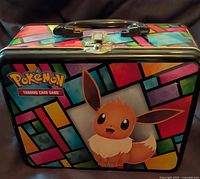 Front view shows Pokémon TCG metal lunch box with Eevee art and multicolor pattern