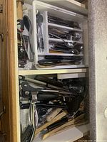 Open kitchen drawers showing flatware organizer filled with knives, forks, spoons and lower drawer with various black-handled utensils and wooden tools
