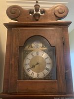 Close view of clock hood showing brass dial, arched glass door, swan-neck pediment, finial