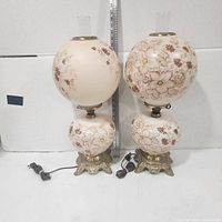 Front view of both matching hand-painted hurricane lamps showing glass globes, bases, chimneys, cords