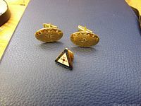 Pair of gold-tone cufflinks and triangular enamel pin displayed on blue surface