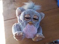 Front view of grey Furby plush showing beak, eyes, pink belly, ears, feet and tufted hair
