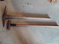 Pickaxe and sledge hammer laid side by side on floor showing full view of handles and heads