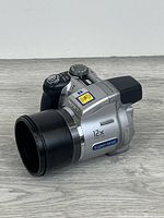 Front angled view showing Sony Cybershot silver body and black 12x zoom lens hood