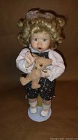 Full standing view of doll on stand holding teddy bear