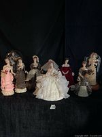 Group shot of eight Victorian style lady figurines arranged in front of two glass cloche domes against black backdrop