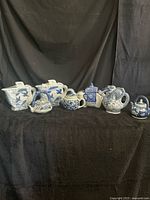 Front view showing all eight blue-and-white ceramic teapots and pitchers lined up on black fabric