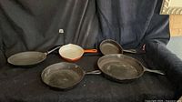 Five pans on black backdrop: three cast iron, one orange enameled cast iron, one carbon steel crepe pan