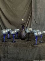 Front view of all ten cobalt blue glass pieces showing decanter with stopper flanked by eight goblets on dark backdrop