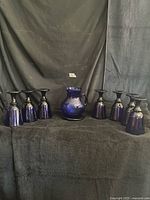 Front view of cobalt blue glass pitcher flanked by seven matching goblets displayed on black fabric backdrop