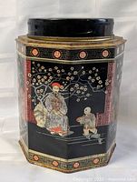 Front view of black hexagonal tea tin showing Asian figural artwork and gold trim