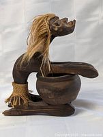 Left side view of carved wooden Tiki figure with straw hair, bowl, and base
