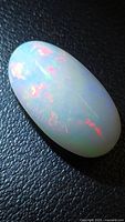 Oval translucent fire opal cabochon on black textured surface displaying red and green flashes