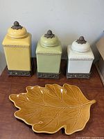 Front view of three ceramic canisters on metal bases and caramel leaf platter