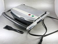Closed panini press with cord and scraper visible