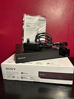 Front view of Sony BDP-S1700U player on box with AC adapter and manual