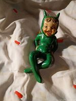 Full view of green devil elf figurine reclining on fabric