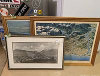 Three framed Vancouver pieces standing together: two colour aerial prints and one black-and-white panoramic photo