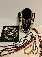 Group shot of all ten necklaces laid out with two on a bust and rest on table and tray