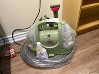 Front view of Bissell Little Green cleaner, coiled clear hose, cleaning formula bottle and pink attachments visible