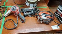 Three corded power tools on table: red heat gun, black/red rotary tool, orange/gray jigsaw