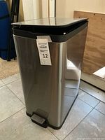 Full view of stainless steel step-pedal trash can showing foot pedal, dent on front, and lid slightly ajar