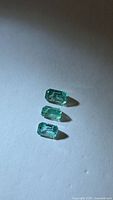 Three emerald-cut emeralds arranged vertically on neutral background