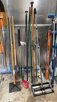 Group of long-handled tools standing upright including post hole digger, spud bar, crowbars, floor scraper, tamper, level, support pole, floor spreader and fork
